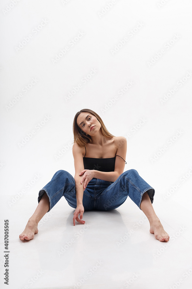 young attractive asian woman with long hair in black top, blue jeans isolated on white studio background. skinny pretty female sitting on cyclorama with bare feet. model tests of beautiful lady