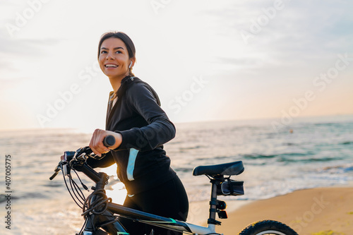 Fototapeta Naklejka Na Ścianę i Meble -  attractive slim woman riding bicycle, sport on morning sunrise beach in fitness suit, healthy lifestyle, listening to music on wireless earphones holding smartphone, relaxing smiling happy