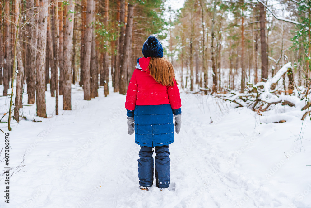 Naklejka premium Rear view of a little girl in a red jacket standing in the middle of a path in a winter pine forest