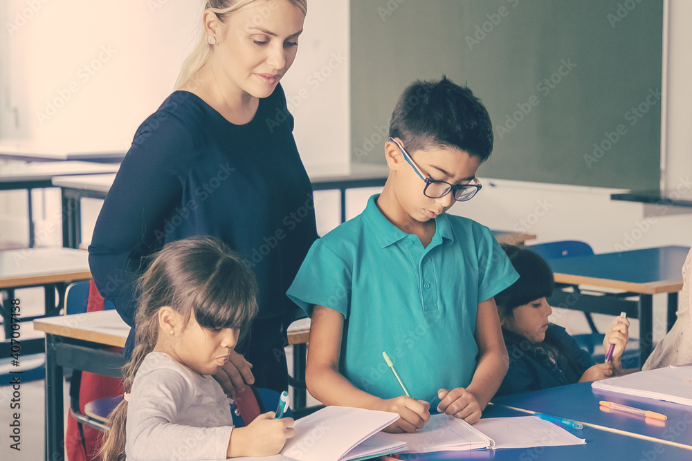 Positive female school teacher watching kids doing their task in class, sitting at desks, drawing and writing in copybooks. Education or back to school concept Positive female school teacher watching kids doing their task in class, sitting at desks, drawing and writing in copybooks. Education or back to school concept
