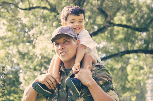 Loving father holding son on neck and walking in city park. Happy Caucasian son sitting on neck of dad in uniform, hugging him and looking away. Family reunion, fatherhood and returning home concept