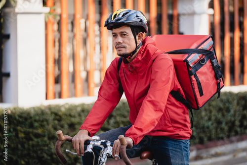 A courier in red uniform with delivery box on back riding bicycle and looking on cellphone to check addres to deliver food to customer. Courier on bicycle delivering food in city. photo