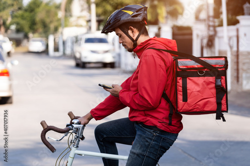 A courier in red uniform with delivery box on back riding bicycle and looking on cellphone to check addres to deliver food to customer. Courier on bicycle delivering food in city.