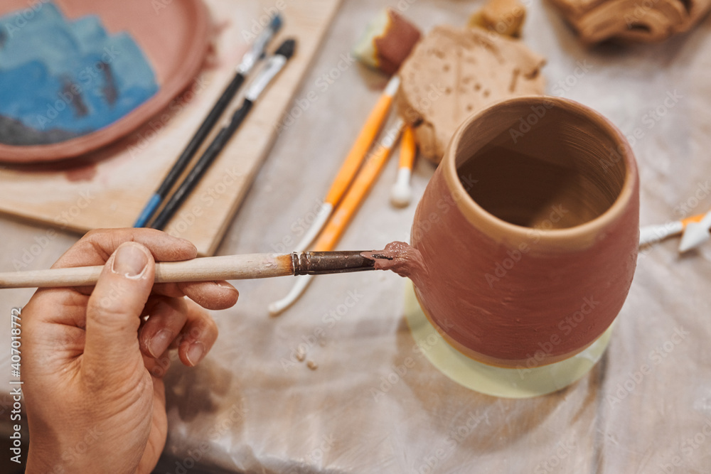 Applying engobe on a ceramic mug with a brush in a pottery workshop. Hands close up