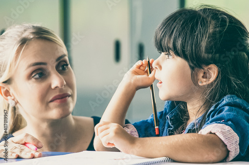 Fototapeta Naklejka Na Ścianę i Meble -  Serious female teacher discussing task with little pupil. Girl sitting at school desk, holding pencil and talking to tutor. Education or back to school concept