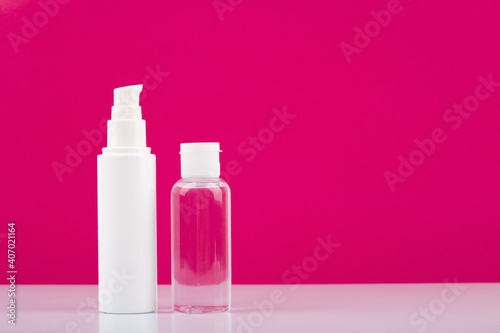 Face cream in white tube and skin lotion in transparent bottle on white table against pink background with copy space