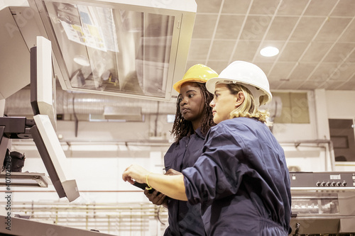 Diverse female industrial workers operating machine on plant floor, looking at monitors over control panel. Side view. Production process or machinery concept