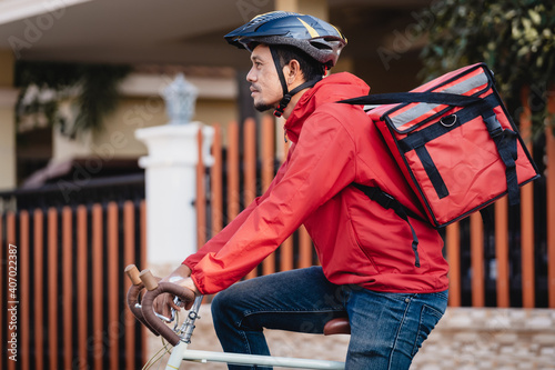 A courier in red uniform with delivery box on back riding bicycle and looking on cellphone to check addres to deliver food to customer. Courier on bicycle delivering food in city. photo