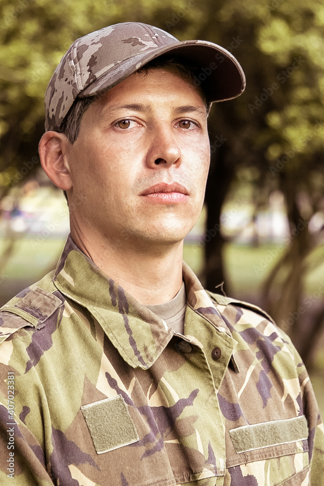 Portrait of serious man in military camouflage uniform standing in park, looking at camera. Green trees in background. Medium shot, low angle. Military man or guard concept