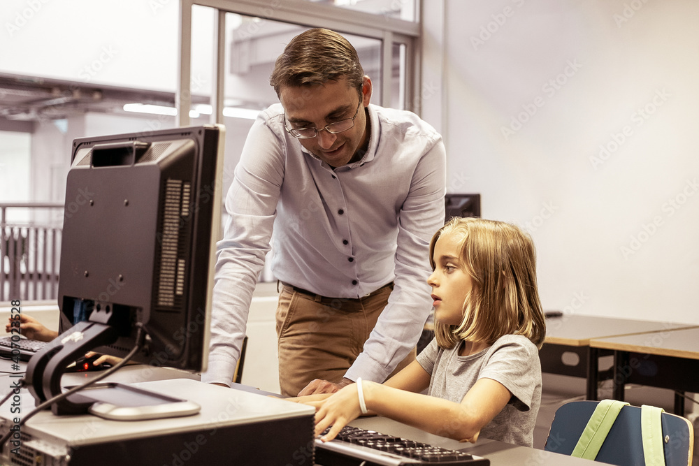 Lovely little blonde student sitting at table during lesson. Caucasian middle-aged lecturer in eyeglasses showing her something, standing and explaining task. Informatics and education concept Lovely little blonde student sitting at table during lesson. Caucasian middle-aged lecturer in eyeglasses showing her something, standing and explaining task. Informatics and education concept