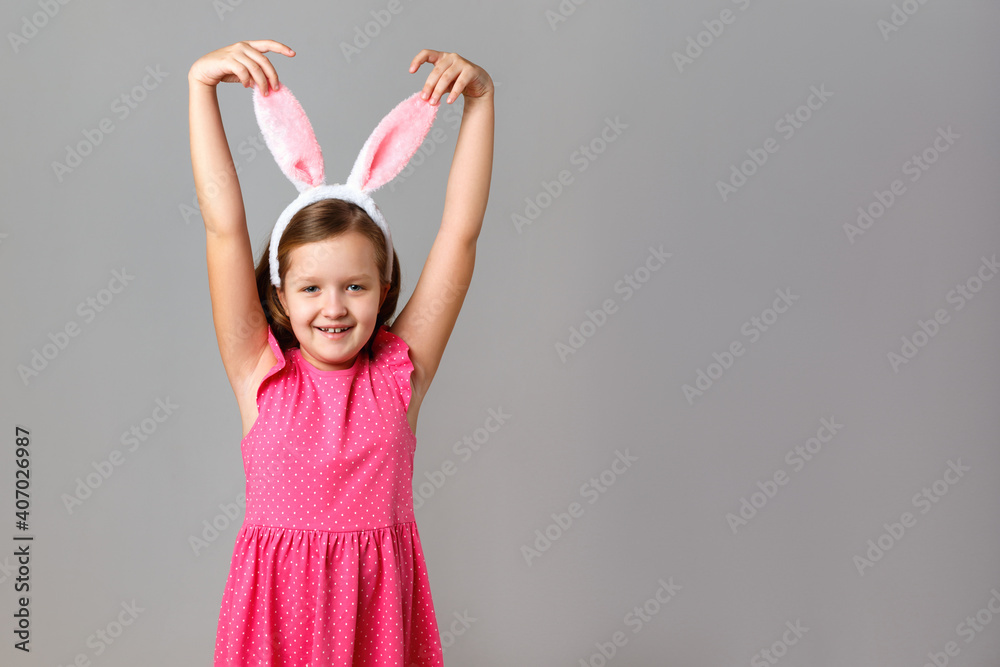 Happy easter. Cheerful little girl in a pink dress with polka dots on a gray background. The child holds his hands behind the rabbit ears. Happy easter. Cheerful little girl in a pink dress with polka dots on a gray background. The child holds his hands behind the rabbit ears.