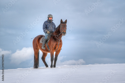 In winter, a lone rider stands on a hill in a snow-covered meadow. In the background there are dark clouds in the sky.