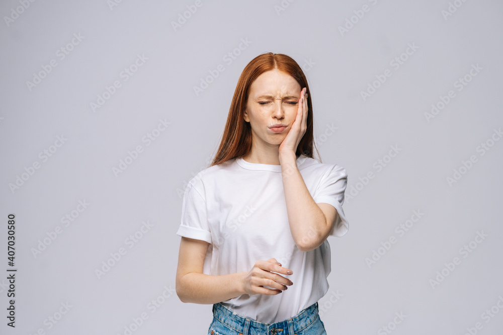 Unhappy upset young woman touches cheek suffering from sudden tooth pain feel on isolated white background. Pretty lady model with red hair emotionally showing facial expressions, copy space