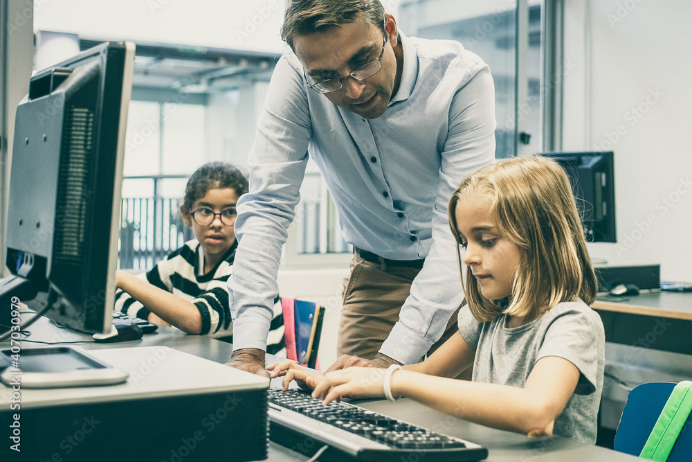 Obraz premium Focused pretty blonde girl typing on keyboard in classroom. Serious middle-aged professor in glasses looking at her, standing near desk and explaining task. Informatics and education concept