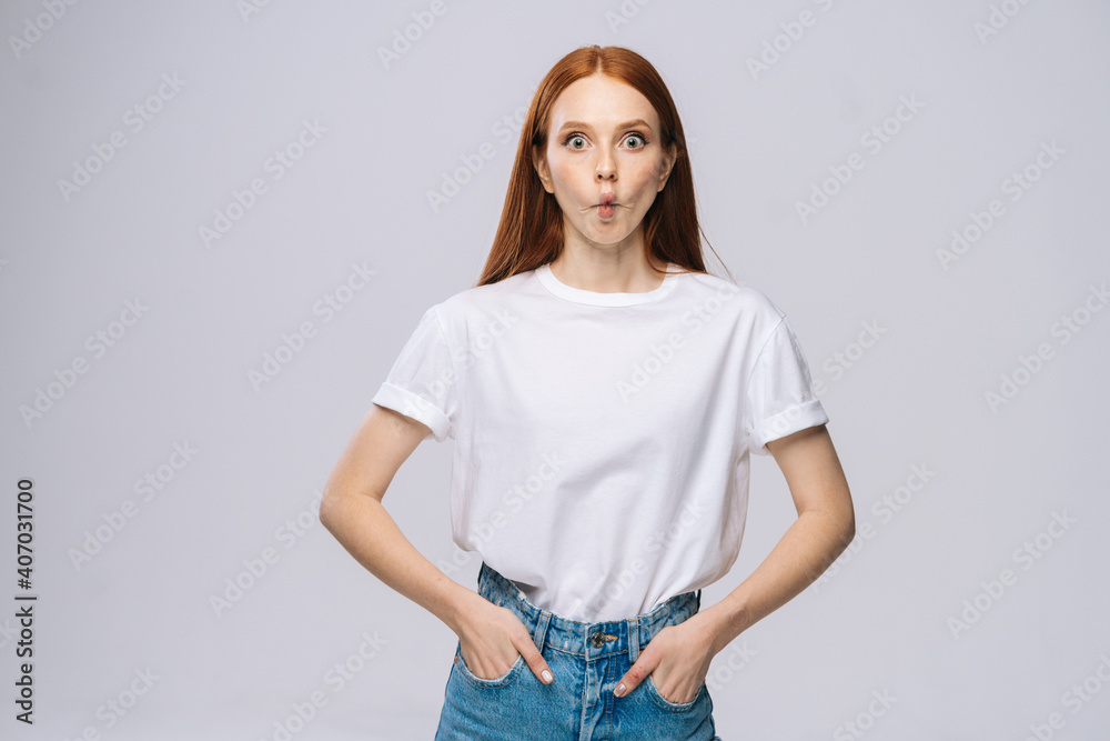 Obraz premium Beautiful young woman wearing T-shirt and denim pants looking at camera with big eyes and fish lip on isolated white background. Pretty lady model with red hair emotionally showing facial expressions.