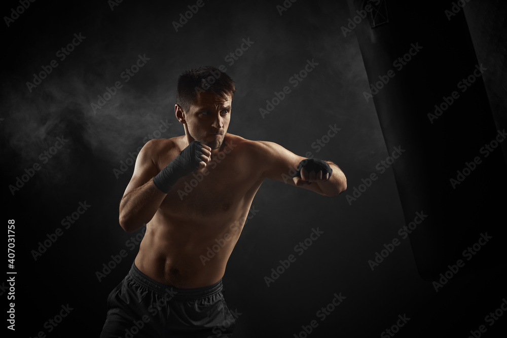 male boxer in black boxing wraps punching in boxing bag on dark background with smoke