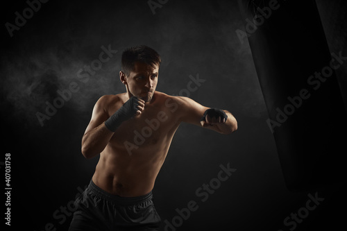 male boxer in black boxing wraps punching in boxing bag on dark background with smoke