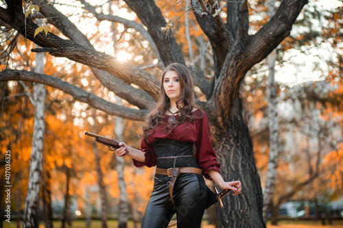 A beautiful girl in a shirt, corset and tight trousers with a saber and a pistol in her hands against the background of an autumn park in the evening. Adventurer woman concept.