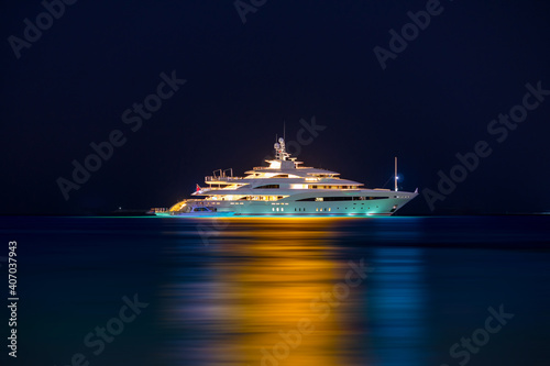 Night view to large illuminated white boat located over horizon, colorful lights coming from yacht reflect on the surface of the the Gulf sea. Shot at blue hour.  