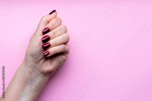 Female hand with beautiful manicure - dark red glittered nails on pink background with copy space