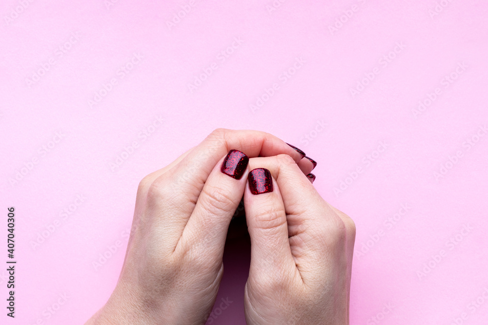 Fototapeta premium Female hands with beautiful manicure - dark red glittered nails on pink background with copy space