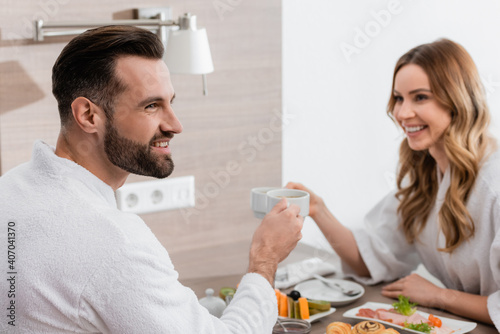 Smiling man in bathrobe holding cup near breakfast and girlfriend on blurred background in hotel