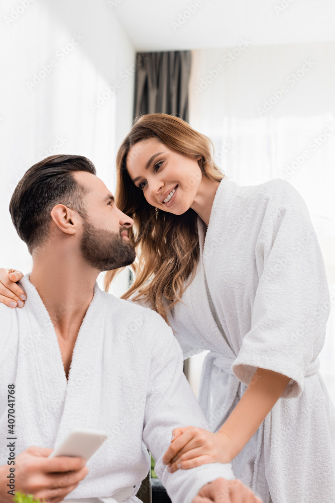 Smiling woman in bathrobe hugging bearded boyfriend with smartphone on blurred foreground in hotel Smiling woman in bathrobe hugging bearded boyfriend with smartphone on blurred foreground in hotel