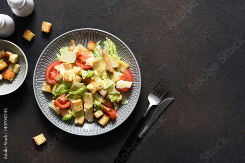 Classic Caesar salad with chicken, tomato and sauce in a plate on a dark concrete background, top view.