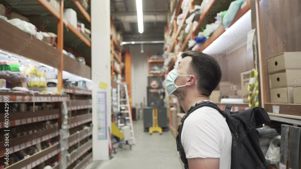 Caucasian man in a hardware store during the ovid19 epidemic in a protective medical mask looking for goods on the top shelves looking at a sample in a mobile phone.Man choosing materials in store. Caucasian man in a hardware store during the ovid19 epidemic in a protective medical mask looking for goods on the top shelves looking at a sample in a mobile phone.Man choosing materials in store.