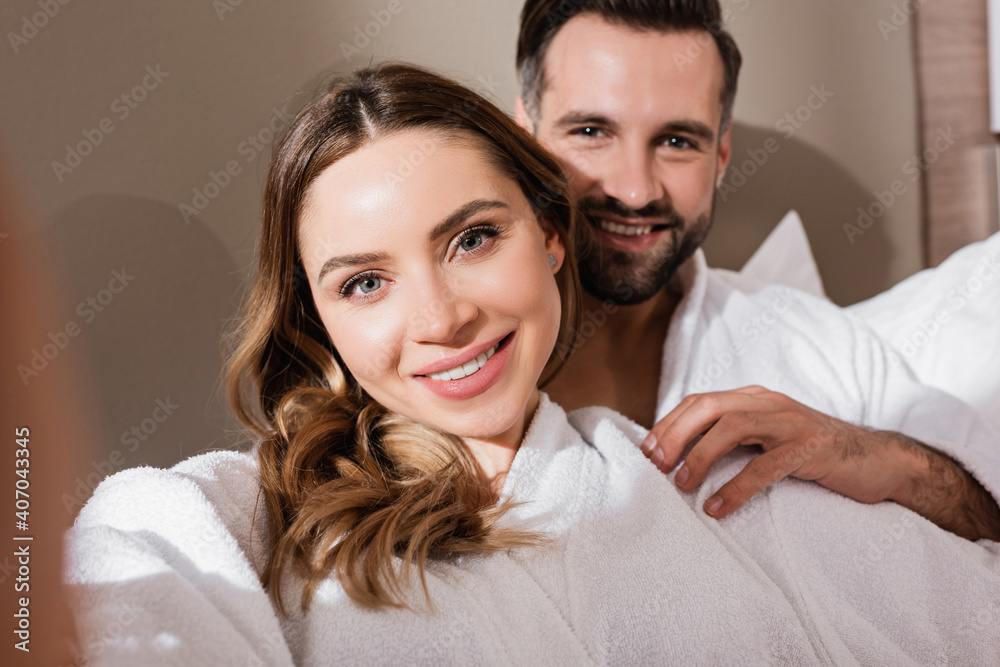 Smiling woman looking at camera near boyfriend in bathrobe on blurred background in hotel Smiling woman looking at camera near boyfriend in bathrobe on blurred background in hotel