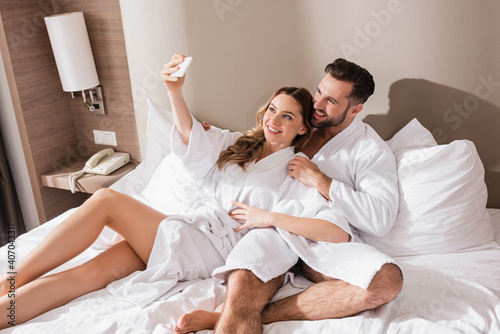 Positive couple in bathrobes taking selfie on smartphone on hotel bed