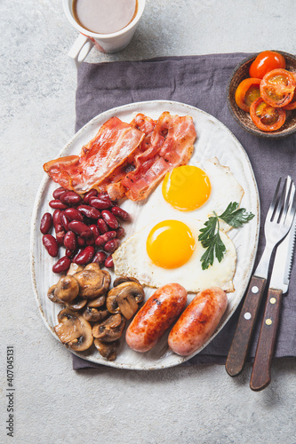 English Breakfast with fried eggs, sausages, bacon, beans, toasts, tomatoes and mushrooms on white plate, concrette background, Top view photo