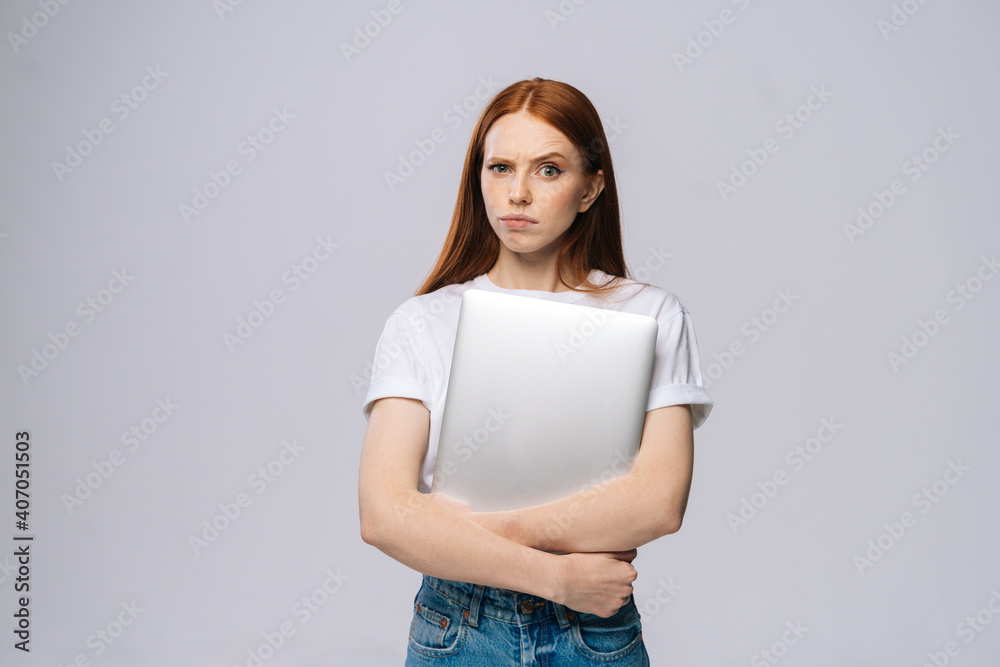 Anxious upset young business woman or student holding laptop computer and looking at camera on isolated gray background. Pretty lady model with red hair emotionally showing facial expressions. Anxious upset young business woman or student holding laptop computer and looking at camera on isolated gray background. Pretty lady model with red hair emotionally showing facial expressions.