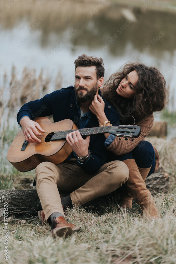 caucasian man is playing guitar with woman by the lake. Young couple is hugging on autumn day outdoors. A bearded man and curly woman in love. Valentine's Day. Concept of love and family.