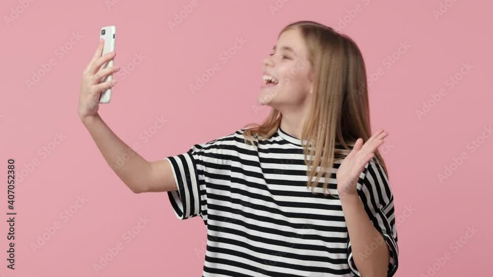 Excited little kid girl 12-13 years old in basic striped t-shirt isolated on pink background studio. Childhood lifestyle concept. Using mobile cell phone talking making video call greeting with hand Excited little kid girl 12-13 years old in basic striped t-shirt isolated on pink background studio. Childhood lifestyle concept. Using mobile cell phone talking making video call greeting with hand