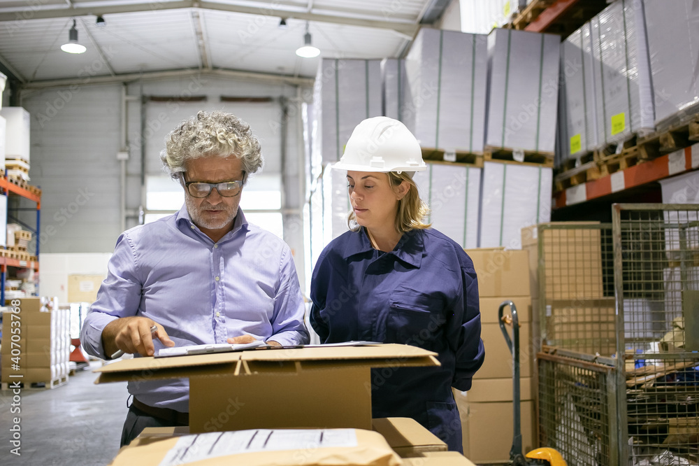 Focused mature inspector consulting female logistic worker while completing documents in warehouse. Medium shot, front view. Labor and inspection concept Focused mature inspector consulting female logistic worker while completing documents in warehouse. Medium shot, front view. Labor and inspection concept