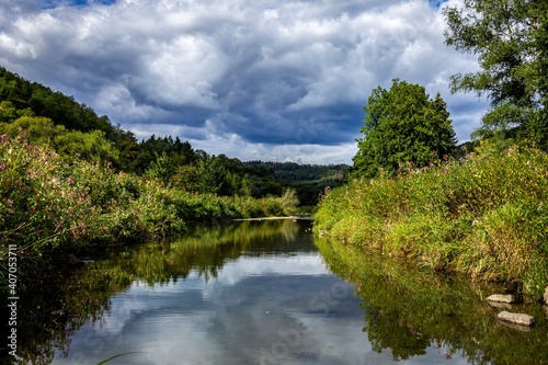 A portrait of a part of the semois river in Vresse-Sur-Semois in the Belgian Ardennes. There is a moody cloudy sky above it, with vegetation growing over the water and it is surrounded by a forest.