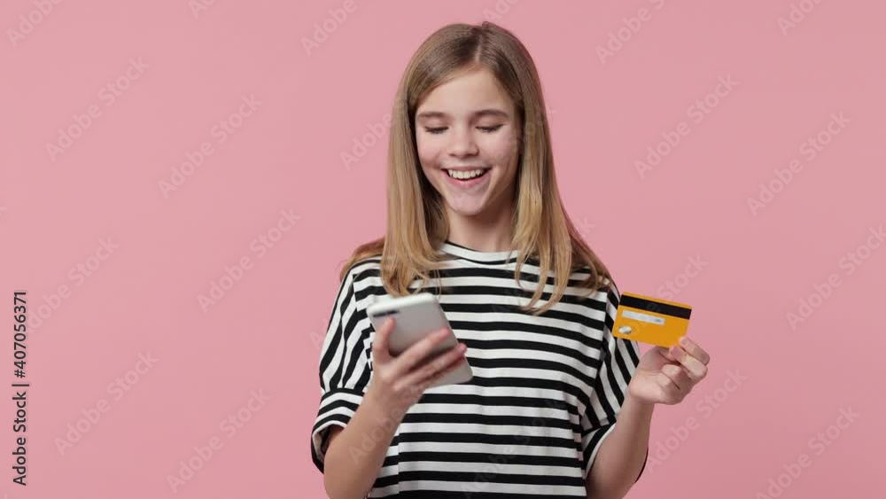 Smiling little blonde kid girl 12-13 years old in basic white black striped t-shirt isolated on pink color background studio. Childhood lifestyle concept. Using mobile cell phone hold credit bank card Smiling little blonde kid girl 12-13 years old in basic white black striped t-shirt isolated on pink color background studio. Childhood lifestyle concept. Using mobile cell phone hold credit bank card