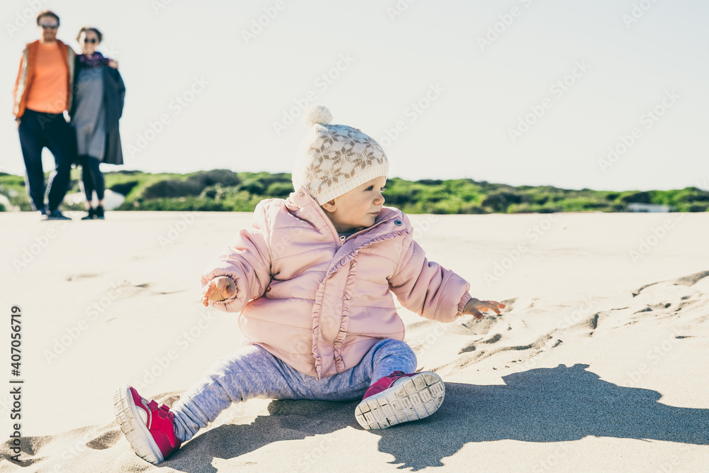 Sweet toddler girl in warm pink jacket playing on sand outdoors. Parents watching little daughter outdoors. Leisure time and parenthood concept