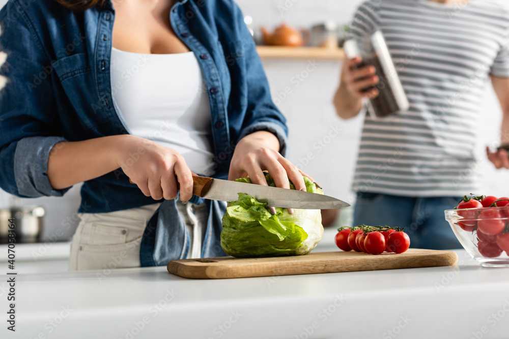 partial view of woman cutting lettuce near ingredients on kitchen table and boyfriend on blurred background