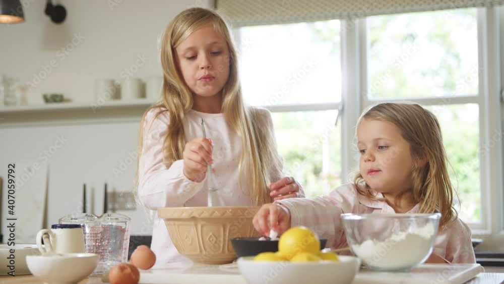 Two girls wearing pyjamas in kitchen mixing ingredients with whisk and adding marshmallows in bowl as they bake at counter together - shot in slow motion Two girls wearing pyjamas in kitchen mixing ingredients with whisk and adding marshmallows in bowl as they bake at counter together - shot in slow motion