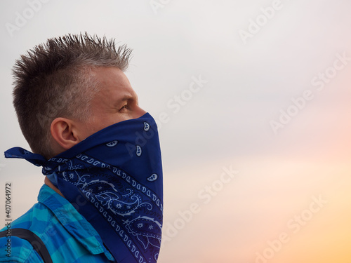 Portrait of a caucasian man in the rays of the setting sun. His face covered with a blue bandana, dressed in a plaid blue shirt. The man has a black backpack. Gray hair styled upwards. photo