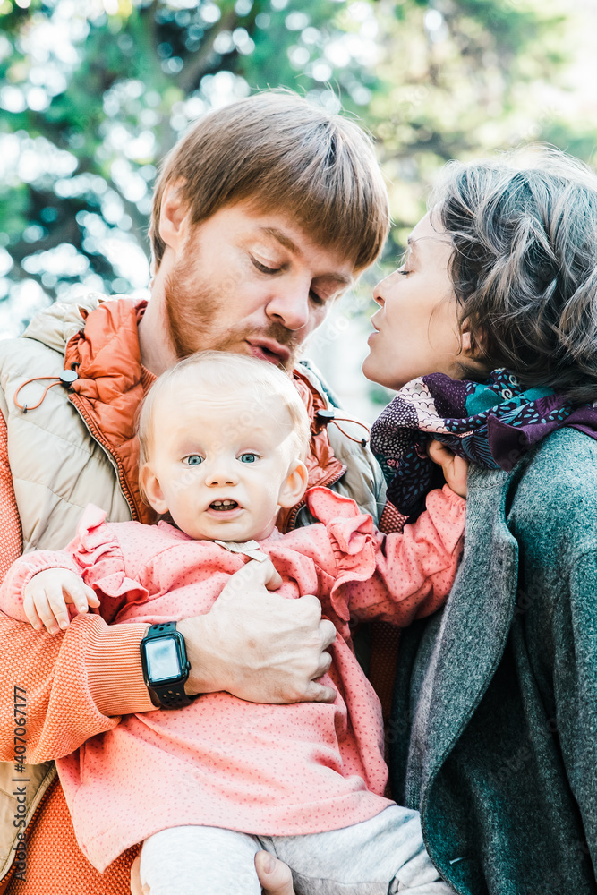 Naklejka premium Cute couple of parents and little daughter posing outdoors. Mom and dad holding cute baby girl in arms, standing in autumn park. Vertical shot. Family portrait concept