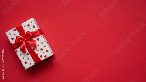 Valentine's day banner concept, gift box wrapped in white paper with red hearts and tied with a red ribbon bow on red background, top view, flat lay, copy space,place for congratulatory text © Komchatnykh Tetiana