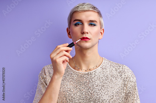 womanlike man is applying lipstick and looking at camera having bright make-up on face, transgender, love cosmetics