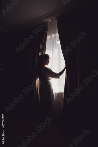 Cute young woman bride in home bathrobe at window in hotel room. Morning of bride on wedding day. Happy female is waiting to meet her groom. Concept of happy and luxury married. Copy space