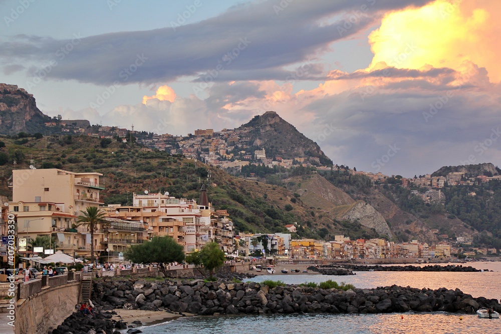 view of a town on a hillside, a town by the sea in Italy, buildings located on hills, light breaking through the clouds, Giardini Naxos