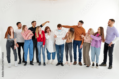 sick male in medical mask stand among healthy people laughing at him, mocking. group of people stand in a row looking at male standing in center, isolated on white studio background