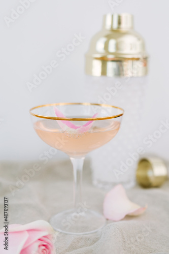 Pink bubbly sparkling cocktail made with champagne, vodka, and rose water, shaken with ice, served in gold rimmed coupe glass, garnished with rose petal. White cocktail shaker in background. photo