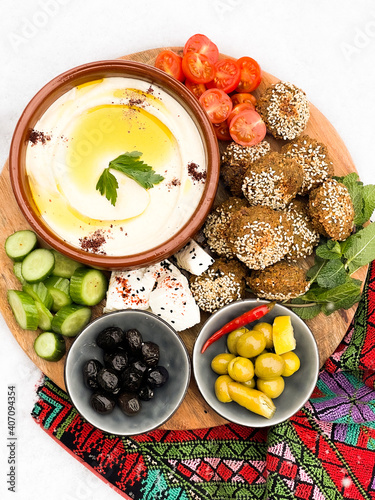 Roasted chickpeas falafel patties with a plate of Hummus, served with fresh vegetables and olives in a wooden plate over light background. Healthy vegan food, clean eating, dieting, top view